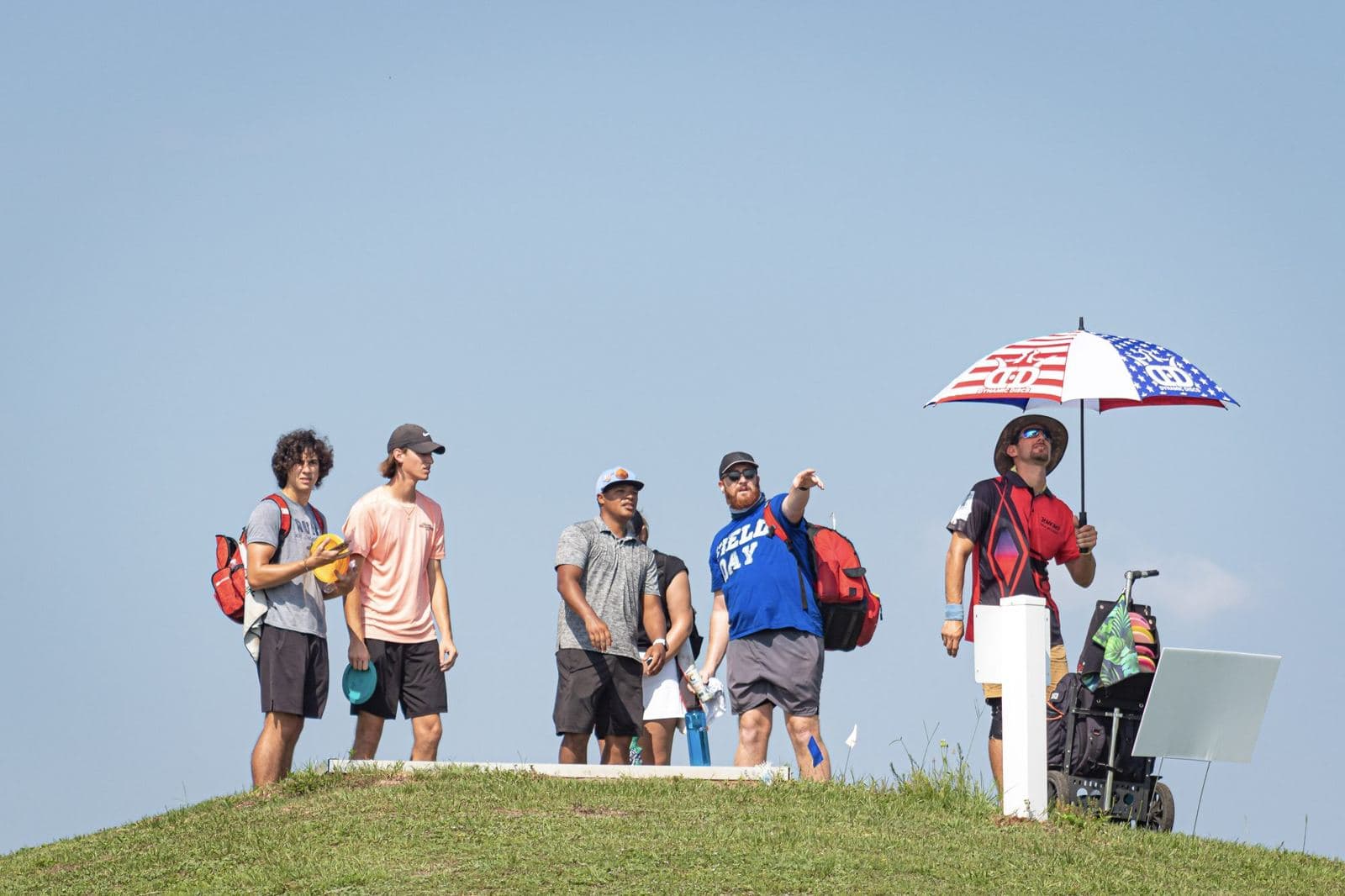 A card of players on a mound under an umbrella, waiting on a tee in July.