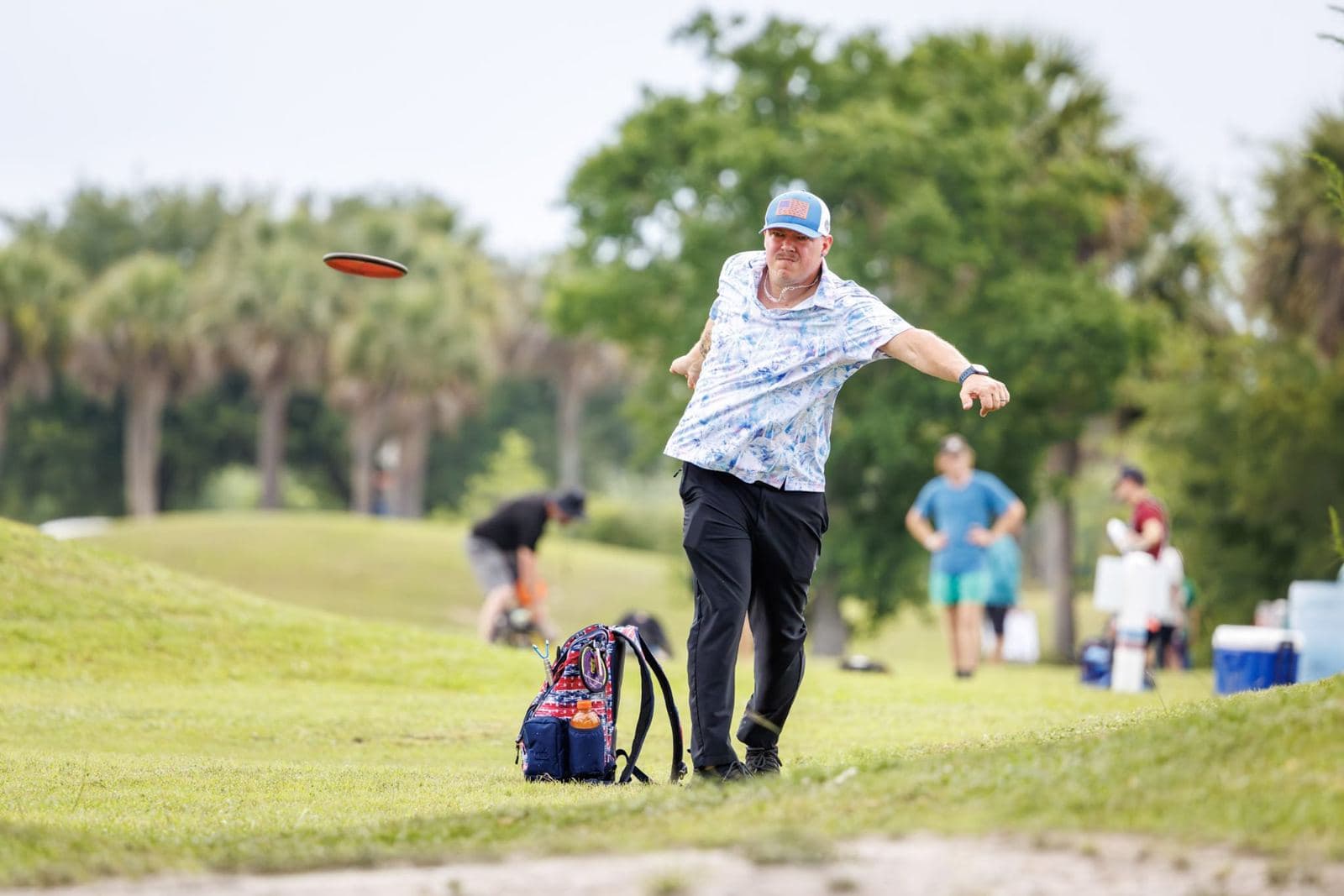 A player in a patterned shirt releases a drive from a grassy rise while a card waits behind him.