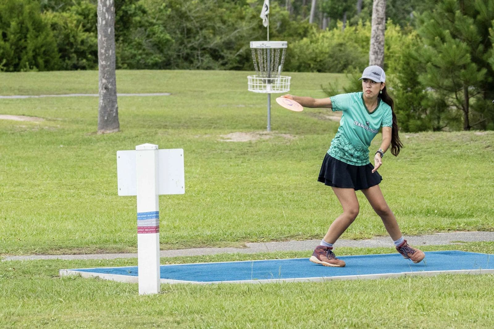 A player throws from a blue tee pad beside a white tee sign, a basket in the distance.